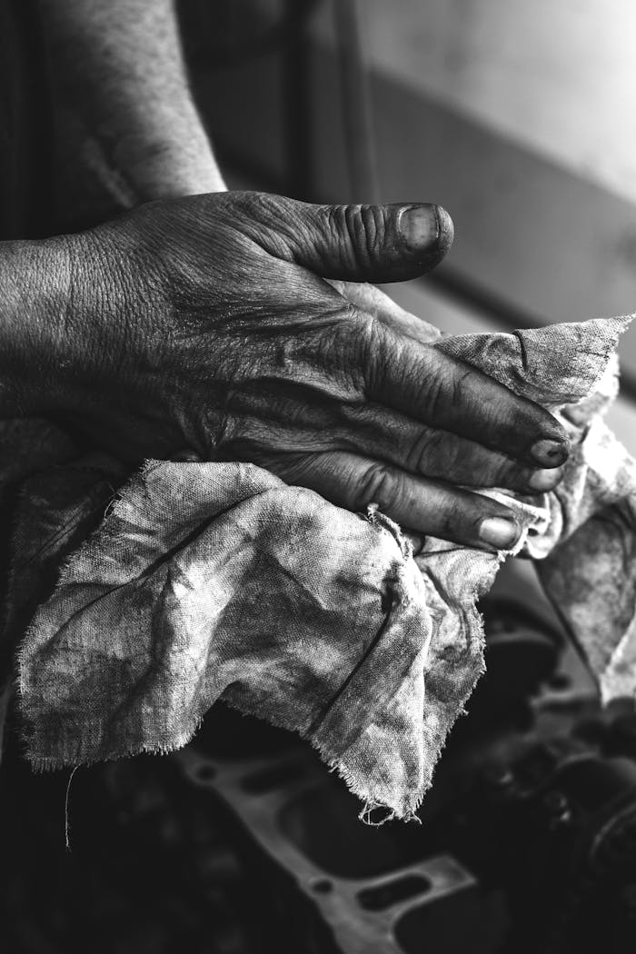 A monochrome image showing dirty hands wiping mechanical parts with a cloth in a workshop setting.